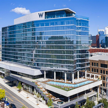 Exterior view of the W Nashville hotel, a modern glass high-rise with a rooftop pool and lounge, set among city streets and surrounding downtown buildings.