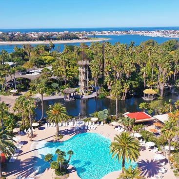Aerial view of a tropical resort with a large outdoor pool surrounded by palm trees and lounge chairs, set beside a lagoon with the ocean and coastline visible in the distance.